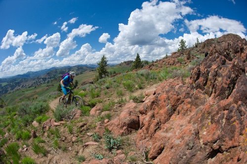 A mountain biker navigates a rocky trail surrounded by green hills and a blue sky with fluffy clouds.