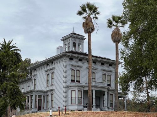 A large, historic gray house with a tower, surrounded by palm trees and a grassy area under a cloudy sky.