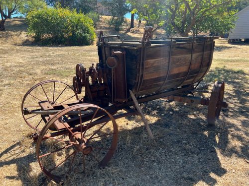 An old wooden cart with metal wheels and machinery, sitting on dry grass under a sunny sky.