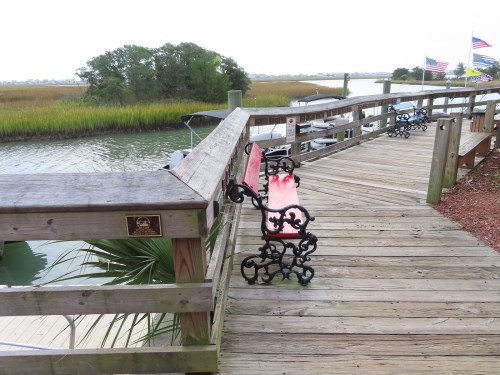 A wooden boardwalk with a red bench overlooking a calm waterway and marshland, with flags in the background.