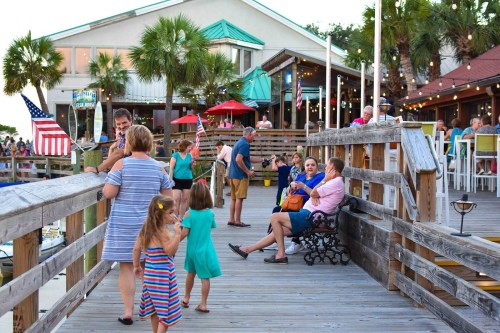 A lively boardwalk scene with families and friends enjoying a waterfront restaurant, surrounded by palm trees and festive decor.