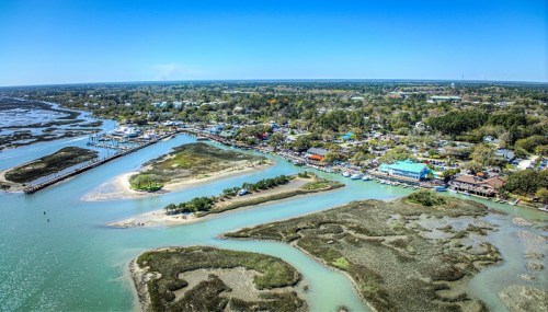 Aerial view of a coastal town with waterways, marshlands, and colorful buildings under a clear blue sky.