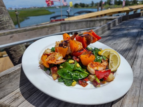 A plate of colorful shrimp and vegetable skewers on a bed of spinach, garnished with lemon, by a waterfront setting.
