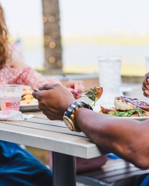 A close-up of hands enjoying a meal outdoors, featuring grilled fish and sides on a table with drinks.