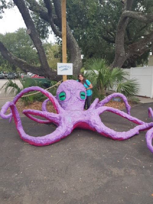A large, purple octopus sculpture with a person standing beside it, surrounded by trees and palm plants.