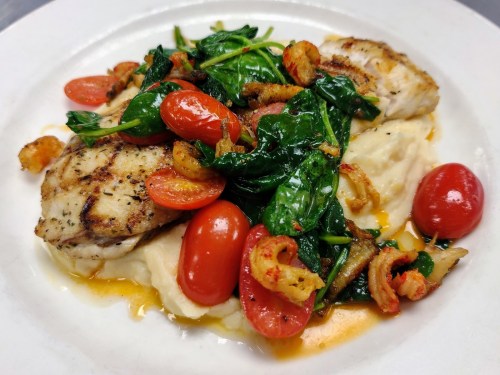 Grilled fish fillets atop creamy mashed potatoes, garnished with sautéed spinach, cherry tomatoes, and crispy bits.