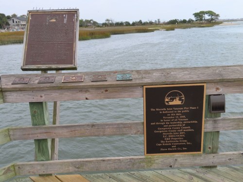 Plaques on a wooden pier overlooking a calm waterway and marshland, commemorating a conservation project.