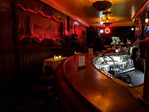 A dimly lit bar with a curved wooden counter, glowing candles, and a well-stocked shelf of drinks in the background.