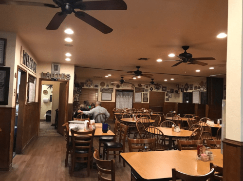 Interior of a cozy restaurant with wooden tables, chairs, ceiling fans, and walls adorned with framed photos.