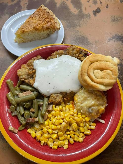 A colorful plate with fried chicken, green beans, corn, cheesy casserole, gravy, and a roll, alongside a slice of cornbread.