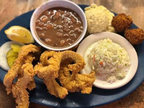 A plate of fried catfish, coleslaw, rice, beans, hushpuppies, and lemon slices, garnished with pickles and onions.