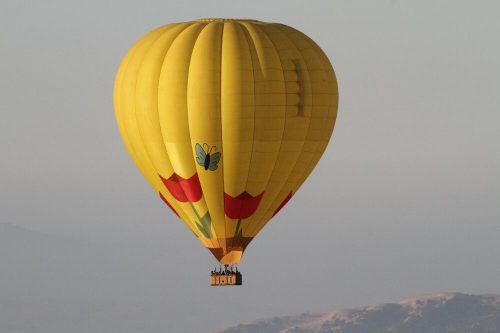 A bright yellow hot air balloon with floral designs floats in the sky, surrounded by mountains in the background.