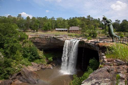 A waterfall cascades over a rocky ledge, with a green statue nearby and lush trees in the background.