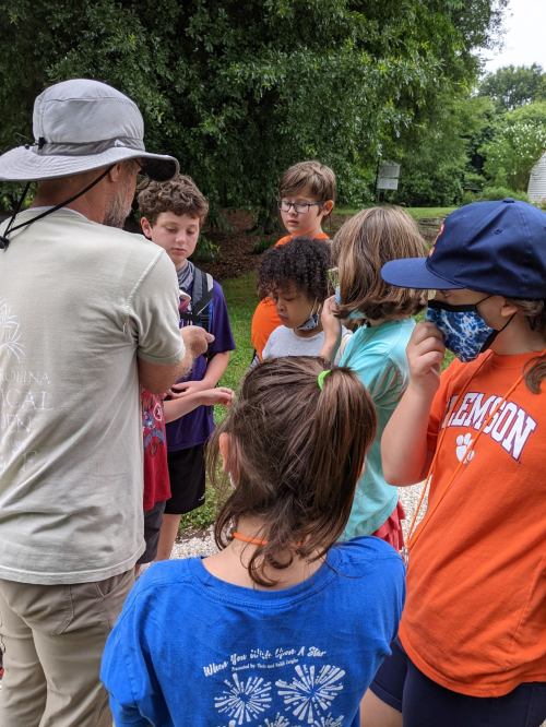 A group of children gathers around an adult, listening intently in a green outdoor setting.