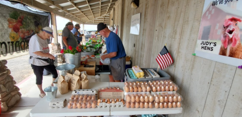 A bustling farmers market with vendors selling eggs, flowers, and produce under a covered area. An American flag is displayed.