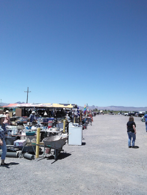 A bustling outdoor market with colorful stalls under a clear blue sky, people browsing and walking along a gravel path.