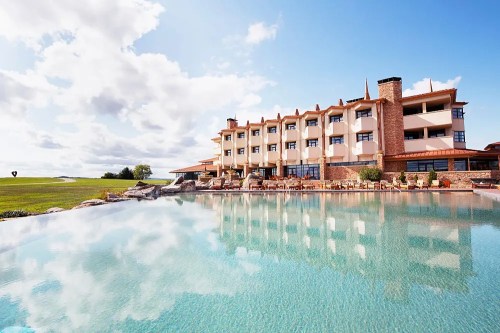 A modern hotel with a stone facade beside a tranquil pool reflecting the sky and clouds.