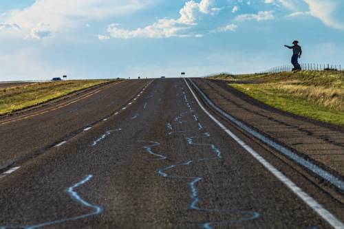 A long, empty road stretches into the distance, with a person standing on the side, waving against a blue sky.