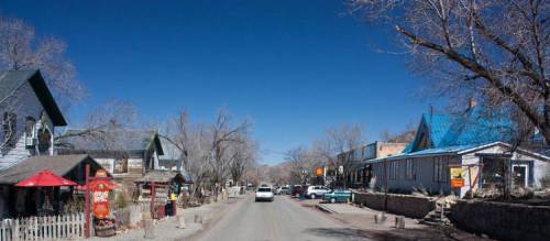 A quiet street lined with quaint buildings and trees under a clear blue sky. Cars are parked along the road.