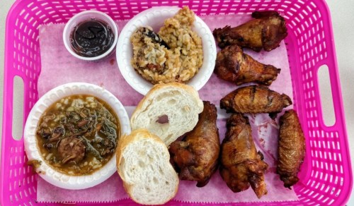 A pink basket filled with fried chicken wings, a bowl of greens, a side of rice, and two pieces of bread.