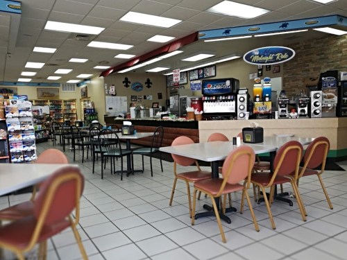 A bright, casual diner interior with tables, chairs, and a counter displaying food and drinks.