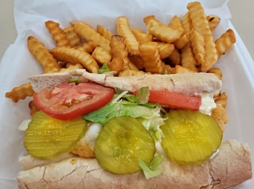 A sandwich with lettuce, tomato, pickles, and chicken, served with crinkle-cut fries on the side.