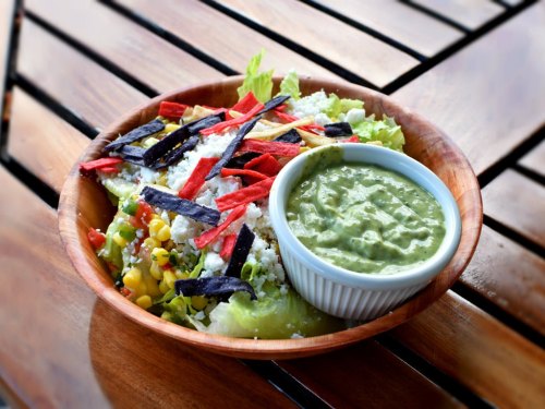 A wooden bowl filled with a colorful salad topped with tortilla strips and a side of green sauce.