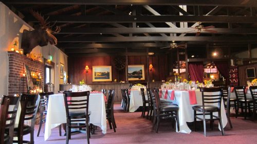 Cozy restaurant interior with wooden beams, tables set for dining, and a moose head mounted on the wall.