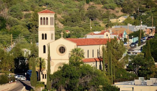 A church with a bell tower surrounded by trees and buildings in a hilly area, under clear blue skies.