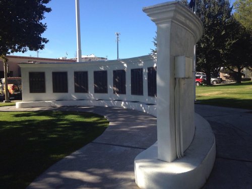 A curved white memorial wall with plaques, set in a park under a clear blue sky.