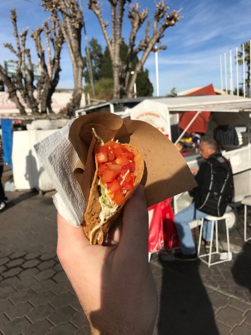 A hand holding a taco filled with tomatoes and lettuce, with food stalls and trees in the background.