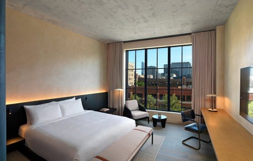 Modern hotel room with a large window, featuring a bed, desk, and seating area, overlooking a cityscape.