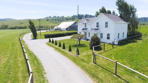 A white farmhouse with a winding driveway, surrounded by green fields and trees under a clear blue sky.