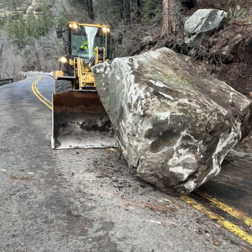 A bulldozer clears a large rock from a winding road surrounded by trees.