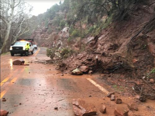 A truck on a muddy road blocked by fallen rocks and debris from a landslide, surrounded by trees and rocky terrain.
