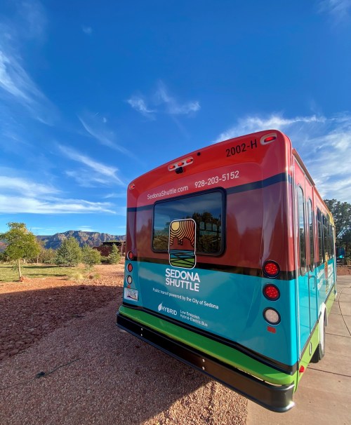 A colorful Sedona Shuttle bus parked outdoors with a scenic backdrop of trees and mountains under a blue sky.