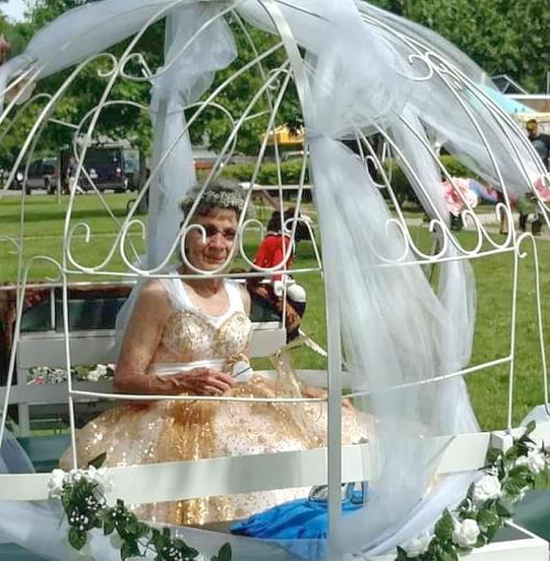 An elderly woman in a sparkling gold dress sits in a decorative birdcage surrounded by greenery and flowers.