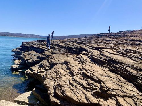 Three people explore rocky terrain by a calm lake under a clear blue sky.
