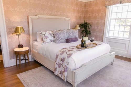 Cozy bedroom with a large bed, decorative pillows, a plant, and a tray with wine on a bedside table. Soft lighting.