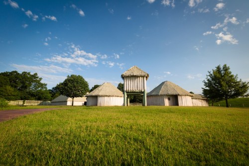 A grassy area featuring traditional thatched-roof structures under a blue sky with scattered clouds.