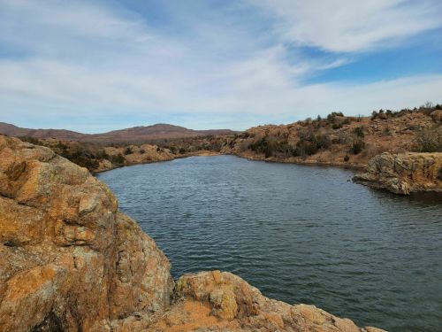 A serene river flows between rocky banks under a blue sky with scattered clouds and distant hills.