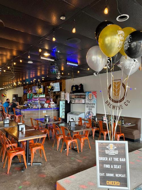 Interior of a lively brewery with festive balloons, tables, and a bar area, inviting patrons to eat and drink.