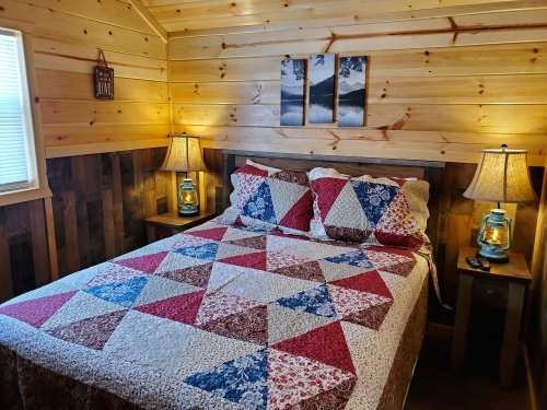 Cozy bedroom with a quilted bedspread, wooden walls, and lamps on bedside tables, featuring nature-themed wall art.