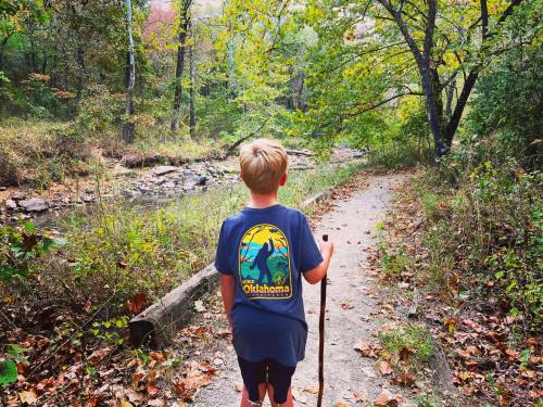 A child with a walking stick stands on a trail surrounded by trees and autumn foliage, facing a creek.