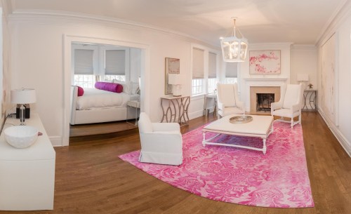 A bright, modern living room with white furniture, a pink rug, and a cozy bedroom visible in the background.
