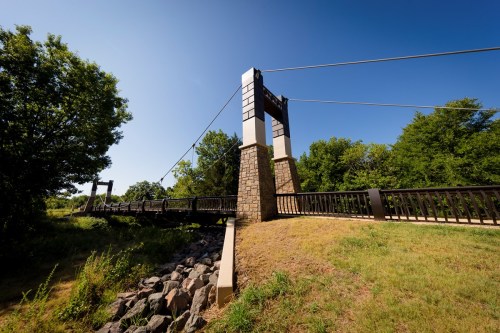 A suspension bridge with stone pillars spans a grassy area, surrounded by trees under a clear blue sky.