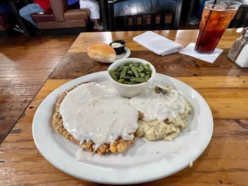 A plate with fried chicken smothered in gravy, mashed potatoes, green beans, and a roll on a wooden table.