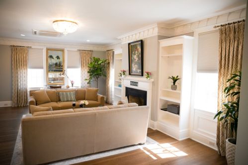 Cozy living room with a beige sofa, fireplace, and large windows, adorned with plants and elegant curtains.