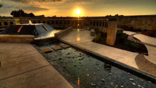 Sunset over a serene courtyard with a fountain and a stream, surrounded by landscaped paths and greenery.