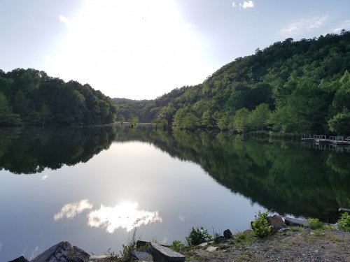 A serene lake surrounded by lush green hills, reflecting the sky and trees in calm waters.